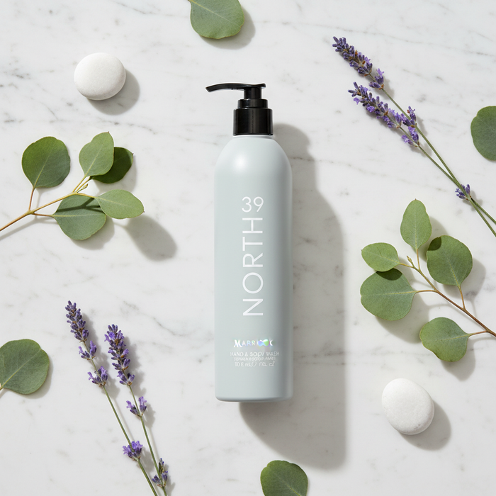 Marriott 39 North Hand & Body Wash 12 oz bottle surrounded by green leaves and purple flowers on marble surface.