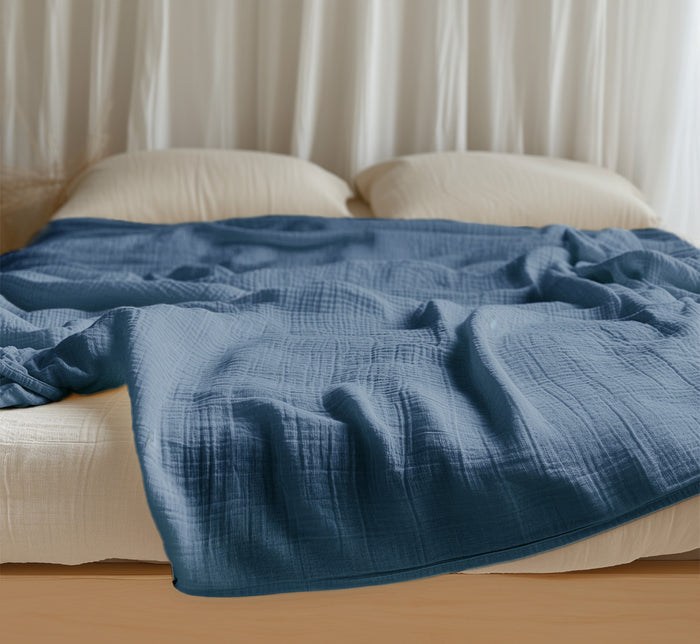 Blue Turmerry organic cotton crinkled reversible muslin blanket on bed with cream pillows and white curtains.