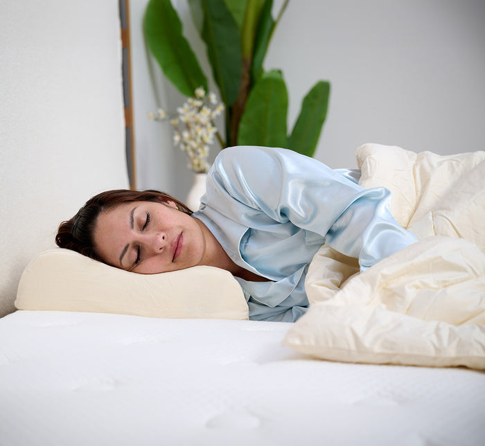 Woman sleeping on her side on Turmerry Latex Contour Pillow wearing light blue silk pajamas, in a bedroom setting.