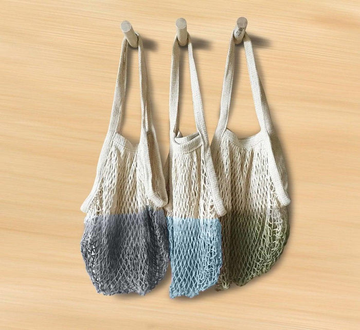 Three Turmerry reusable grocery shopping bags with mesh design in grey, blue, and green hues, hanging on wooden pegs.