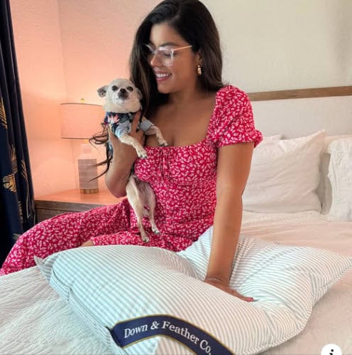 Woman in a pink dress holding a small dog on a bed with Down & Feather Co. branding.